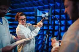 Three people wearing white lab coats and holding space testing equipment stand in a room full of blue foam cones.